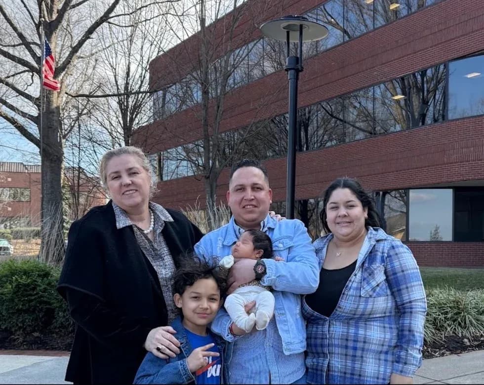 A family of five, including a father holding a newborn baby, poses outside an office building with an American flag flying nearby.