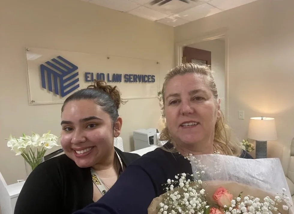 Two women smile for a selfie in the office in front of the Eijo Law Services sign, one holding a bouquet of flowers wrapped in paper.