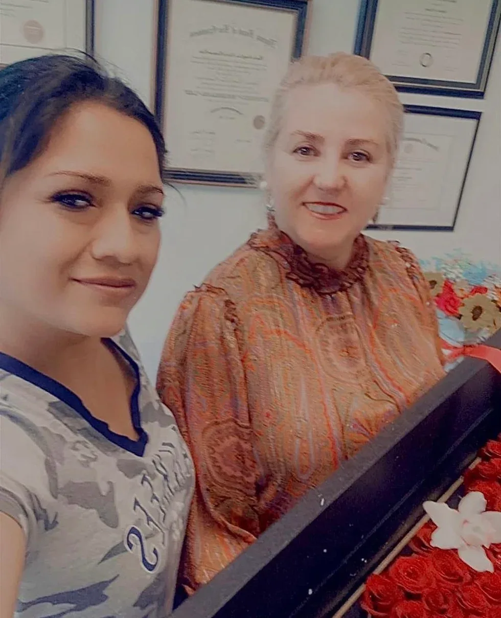 A young woman takes a selfie with Maria Eijo in the office, with the firm’s framed certificates on the wall and a box of red roses in the foreground.
